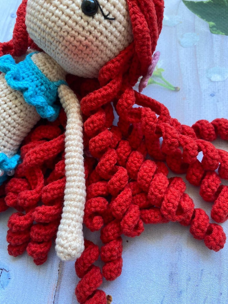 May include: A crocheted doll with long, curly red hair and a blue top. The doll is made with white yarn and has a sweet, innocent expression.