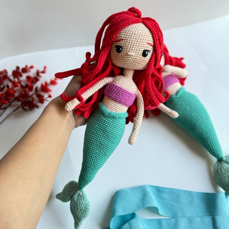 May include: A crocheted mermaid doll with red hair, a green tail, and a purple top. The doll is holding a blue ribbon.