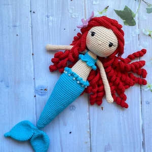 May include: A crocheted mermaid doll with a blue tail, red hair, and a pink flower on her head.