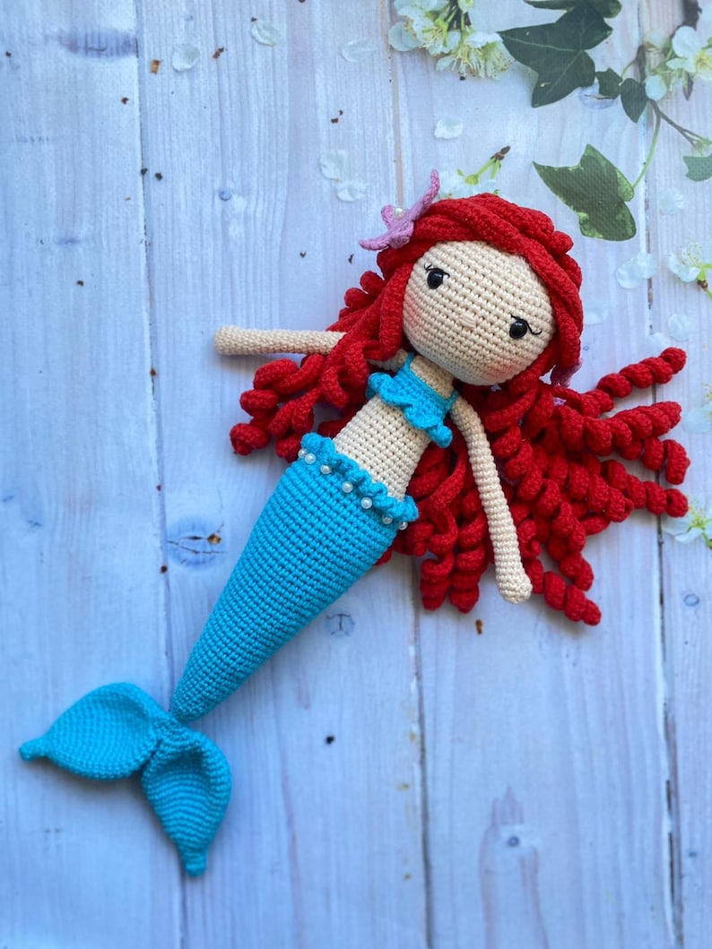 May include: A crocheted mermaid doll with a blue tail, red hair, and a pink flower on her head.