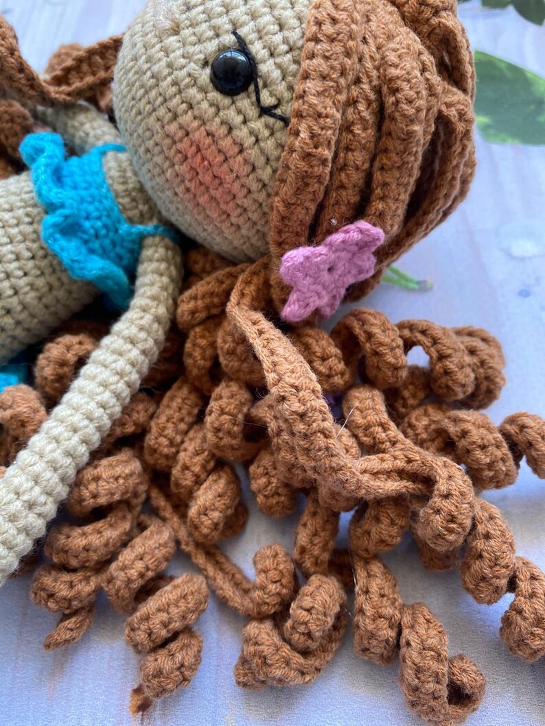 May include: A crocheted doll with brown hair, a blue top, and a pink flower in her hair.