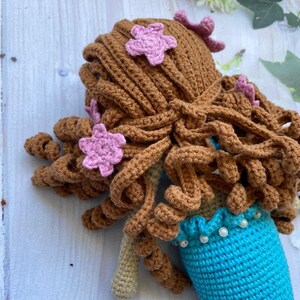 May include: A crocheted mermaid doll with brown hair and pink flower accents. The doll is wearing a turquoise tail with a white pearl trim.