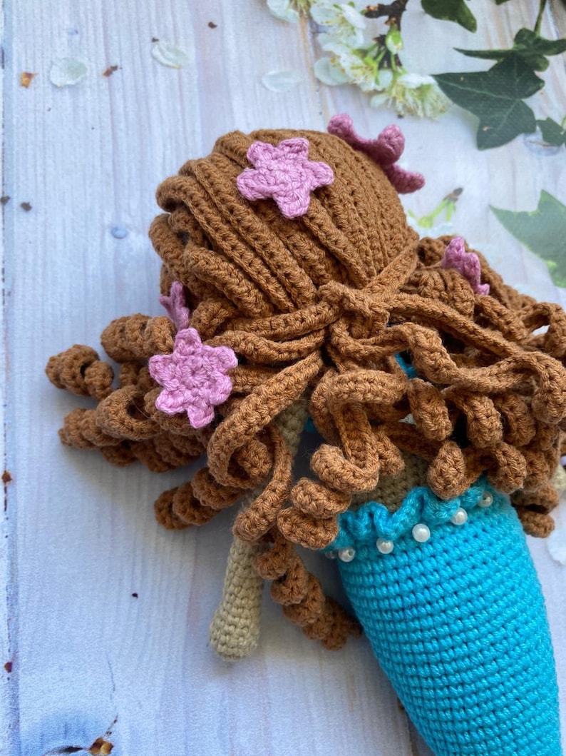 May include: A crocheted mermaid doll with brown hair and pink flower accents. The doll is wearing a turquoise tail with a white pearl trim.
