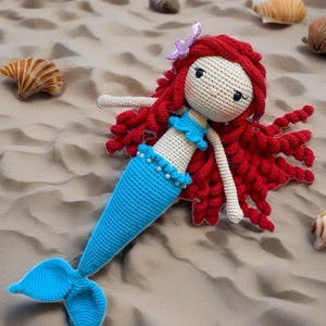 May include: A crocheted mermaid doll with red curly hair, a blue tail, and a white top. The doll is lying on a sandy beach.