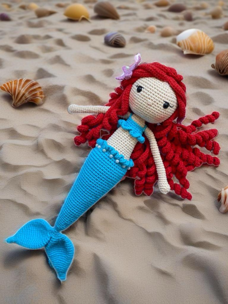 May include: A crocheted mermaid doll with red curly hair, a blue tail, and a white top. The doll is lying on a sandy beach.