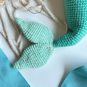 May include: A crocheted mermaid tail with a light green and dark green ombre color scheme. The tail is made with a fine yarn and has a delicate, detailed design.