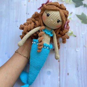 May include: A crocheted mermaid doll with brown hair, a blue tail, and a pink flower in her hair.
