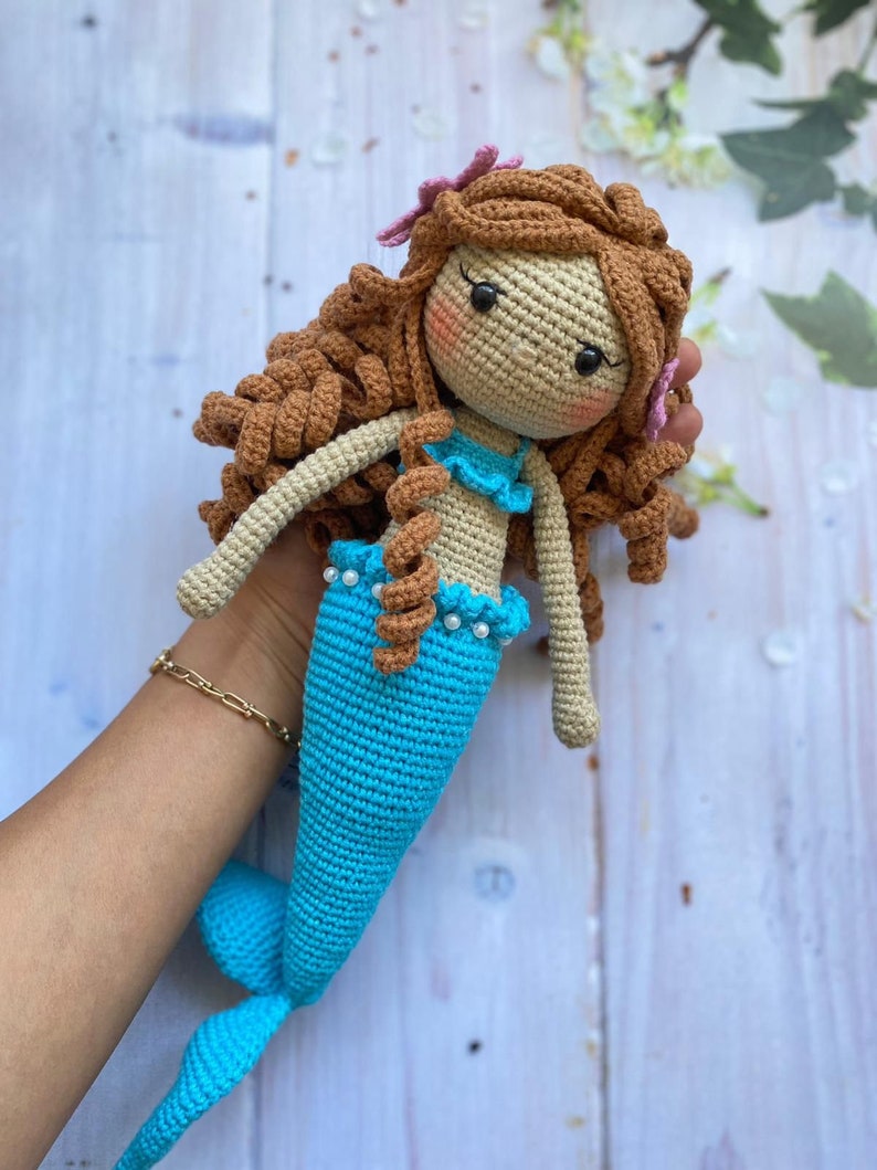 May include: A crocheted mermaid doll with brown hair, a blue tail, and a pink flower in her hair.