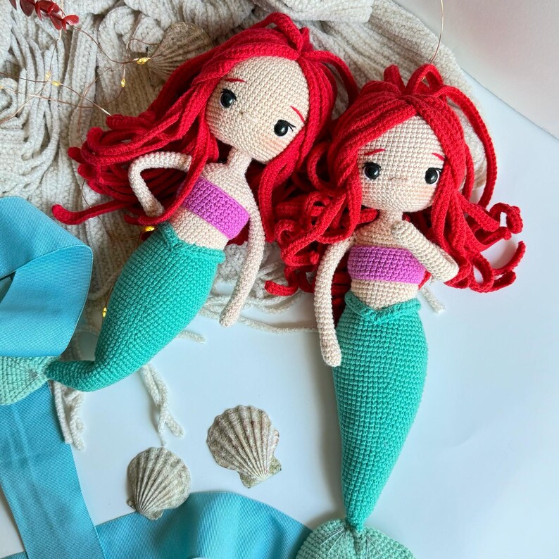 May include: Two crocheted mermaid dolls with red hair and teal tails. The dolls are wearing pink and teal tops.