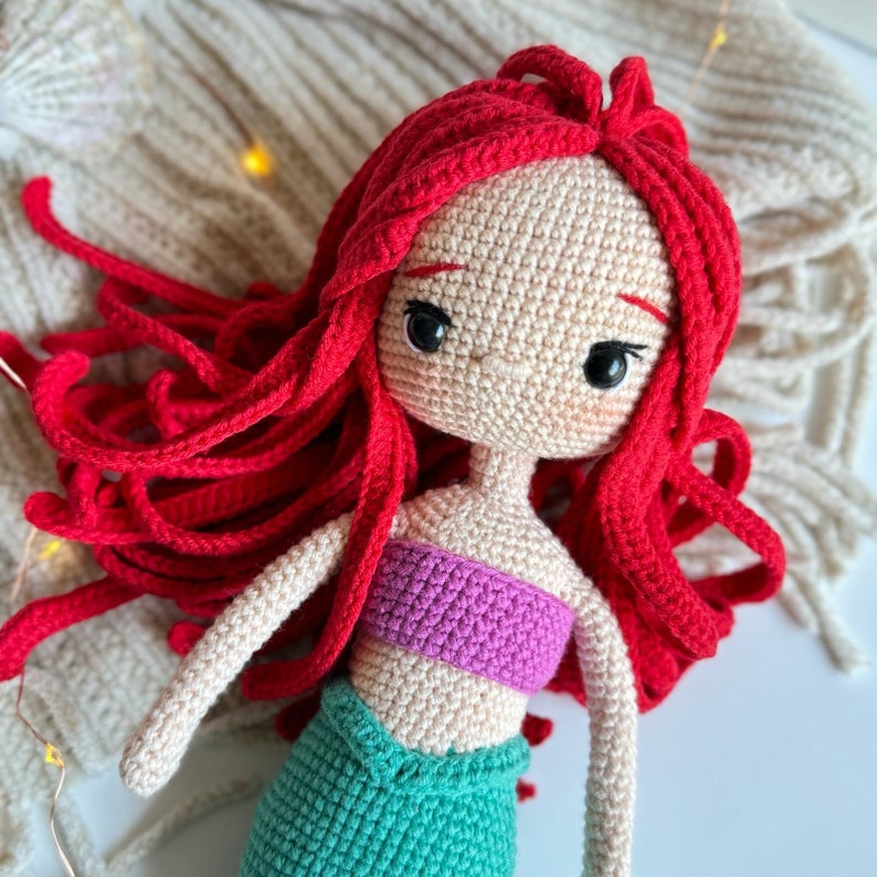 May include: A crocheted doll with long red hair, a pink top, and a green mermaid tail. The doll has a sweet expression with large brown eyes.