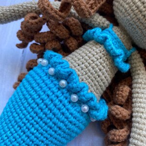 May include: A crocheted mermaid doll with brown hair, a beige top, and a blue tail. The doll is wearing a white pearl trim around the waist.