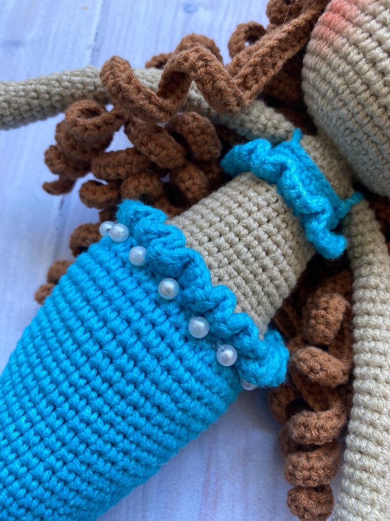May include: A crocheted mermaid doll with brown hair, a beige top, and a blue tail. The doll is wearing a white pearl trim around the waist.
