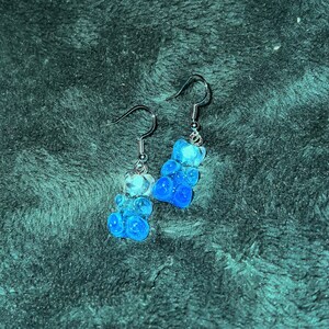 May include: A pair of dangle earrings featuring blue and clear gummy bear charms. The charms are ombre, fading from clear to blue.