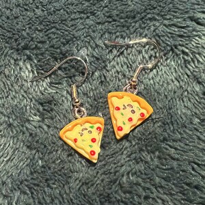 May include: A pair of silver dangle earrings featuring a miniature pizza slice charm. The pizza slice is yellow with red and green toppings.