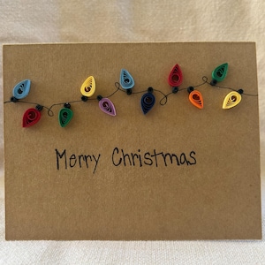 Puede incluir: Una tarjeta de Navidad hecha a mano con un fondo de papel marrón. La tarjeta presenta una cadena de luces navideñas de papel quilling de colores. Las palabras "Merry Christmas" están escritas a mano con tinta negra debajo.