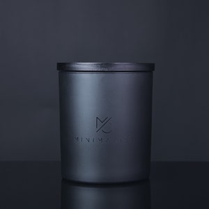 Könnte beinhalten: Ein schwarzer zylindrischer Kerzenbehälter mit schwarzem Deckel. Der Behälter hat ein minimalistisches Logo mit dem Text "MINIMALIST" auf der Vorderseite.