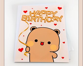 May include: A greeting card with a cartoon bear holding a gold ribbon. The words "Happy Birthday" are in gold lettering above the bear. Red hearts are scattered around the card.