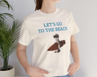 Puede incluir: Camiseta blanca con un gráfico azul de un gato con gafas de sol y sosteniendo una tabla de surf. El texto "Let's go to the beach" está impreso en azul en la camiseta.