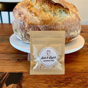 May include: A loaf of sourdough bread on a white cake stand with a small brown paper bag containing sourdough starter. The bag has a white sticker with the text "Salt & Lightt Sourdough Goods" and a drawing of a jar.