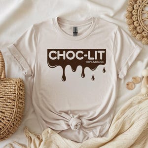 May include: Beige t-shirt with the text "CHOC-LIT 100% Melanin" in a brown, dripping font. The shirt is knotted at the waist and is displayed on a neutral-toned surface with decorative items.