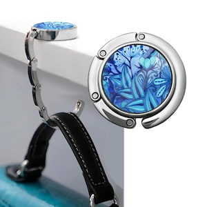 May include: A silver purse hook with a blue floral design. The hook is attached to a black purse handle. The hook is designed to hang a purse from a table or other surface.