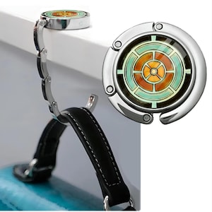 May include: A silver purse hook with a decorative circular top featuring a geometric design in shades of orange, green, and brown. The hook is attached to a chain and is holding a black purse handle. The hook is designed to hang a purse from a table.