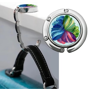 May include: A silver-toned, foldable hook with a colorful abstract design. The hook is closed and ready to be used. It is a handy accessory for hanging bags, purses, or other items.