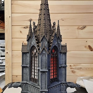 Op de afbeelding: Een gedetailleerd miniatuur gotisch kerkmodel. De structuur is voornamelijk grijs met bruine accenten, met ingewikkelde architectonische details, gewelfde ramen en een kruis op de top. Waterspuwers sieren de buitenkant.