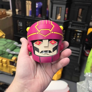 Op de afbeelding: Een magenta Sentinel robot hoofd figuur met gele accenten. Het robothoofd heeft een vierkant gezicht met rechthoekige oogdetails en een witte kin. De figuur wordt in een hand gehouden.