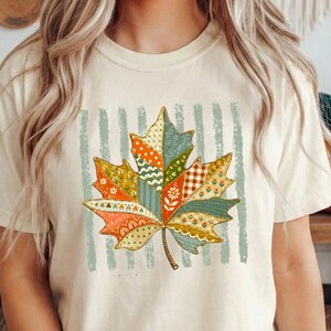 May include: An ivory short-sleeve t-shirt featuring a large central graphic of a maple leaf composed of various colourful patchwork patterns in shades of orange, green, and yellow, set against a background of vertical light green stripes.