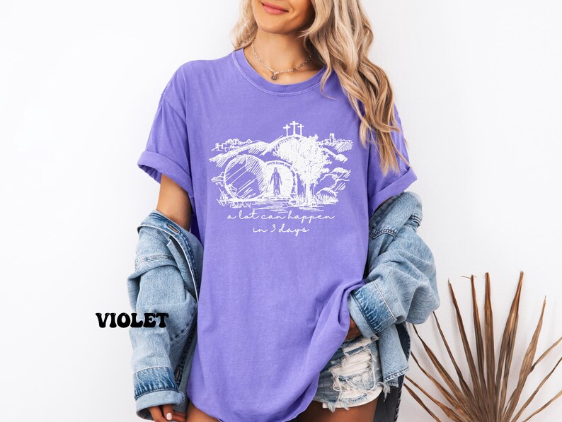 May include: Purple t-shirt with a white graphic of a person walking through a stone archway with a cross above. The text "a lot can happen in 3 days" is below the graphic.
