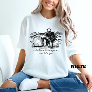 May include: White t-shirt with a black and white illustration of a person walking towards a tomb with a cross above it. The text "a lot can happen in 3 days" is printed below the illustration.