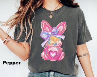 Peut inclure: Un t-shirt gris foncé avec un lapin de dessin animé coloré portant un nœud violet et bleu. Le lapin tient un cœur rose.