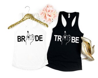 May include: Two tank tops, one white and one black, with the words "BRIDE" and "TRIBE" printed on them. Each top features a hand with a ring on the ring finger, giving the middle finger. A gold sequined hanger and a pair of gold heels are also visible.
