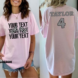 Pu&ograve; includere: Maglietta rosa con un designo a scacchi e il testo "YOUR TEXT" sul davanti. La parte posteriore della maglietta ha il testo "TAYLOR" e il numero "4" in un designo a scacchi.