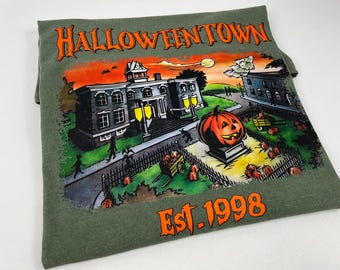 May include: Green t-shirt with a Halloween-themed graphic. The design features a spooky town scene with a jack-o'-lantern, buildings, and the text "Halloweentown Est. 1998" in orange. The shirt is made of a soft material.