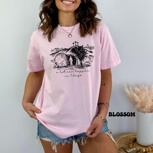 May include: A pink t-shirt with a black graphic of a tomb, a cross, and a figure walking away from the tomb. The text "a lot can happen in 3 days" is printed below the graphic.