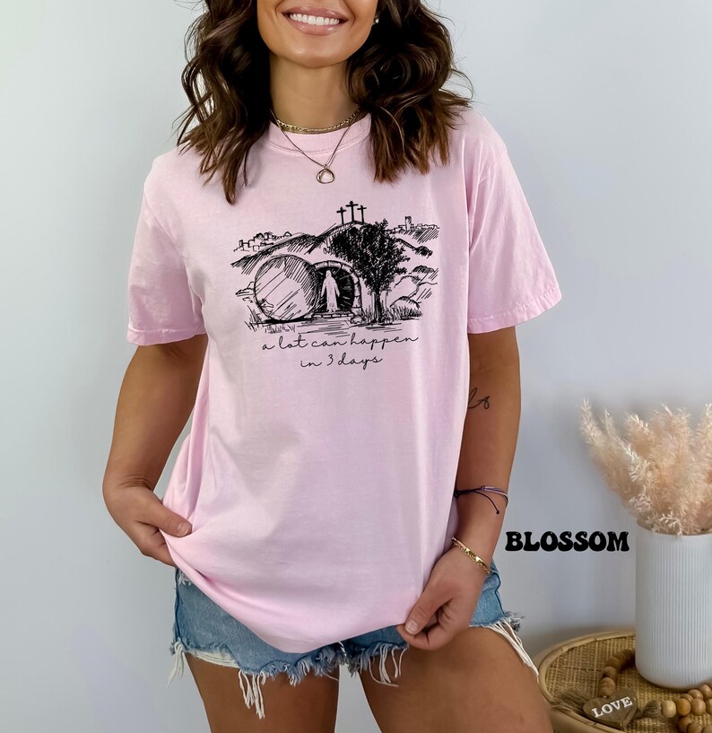 May include: A pink t-shirt with a black graphic of a tomb, a cross, and a figure walking away from the tomb. The text "a lot can happen in 3 days" is printed below the graphic.