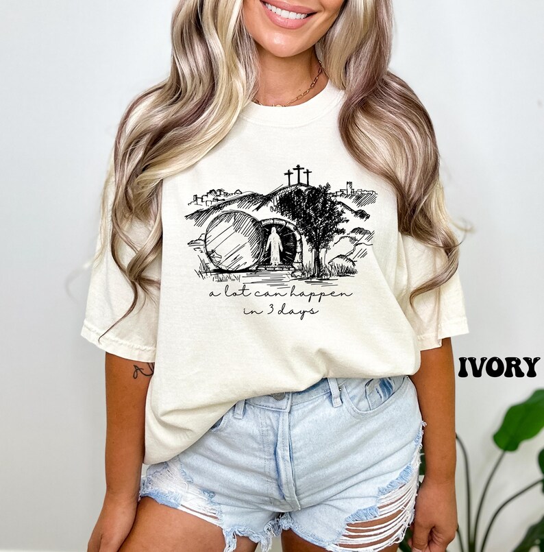 May include: A white t-shirt with a black and white illustration of a tomb, a figure walking away from it, and the text "a lot can happen in 3 days".