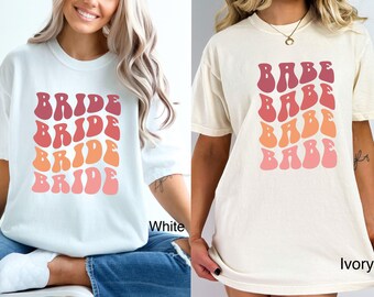 May include: Two t-shirts, one white and one ivory, each with a retro-style graphic. The white shirt features the word "BRIDE" stacked four times in shades of red and orange. The ivory shirt displays the word "BABE" in a similar design.