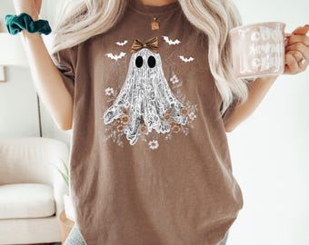 May include: Brown t-shirt with a ghost design, bow, floral details, and bats. The ghost has black eyes and a lace-like outfit. The shirt is a relaxed fit, suitable for everyday wear. The design has a Halloween theme.