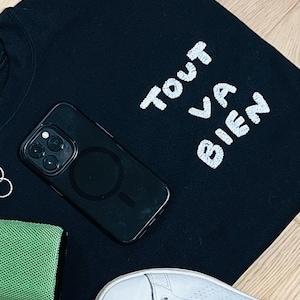 May include: A black sweatshirt featuring the white embroidered text "TOUT VA BIEN" in a casual font. The garment is displayed alongside a black smartphone with a circular magnetic charging ring on the back, a green textured object, and white sneakers.