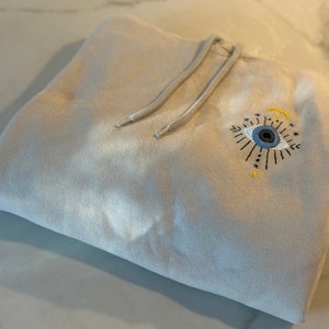 May include: A beige hoodie with a blue and yellow embroidered eye design. The eye has a crescent moon and stars around it.