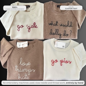 May include: Four crewneck sweaters in cream and tan colors, each featuring hand-embroidered text. The cream sweaters display 'go 'gate' and 'go pios' in red thread. The tan sweaters feature 'what would dolly do?' and 'love trumps hate'.