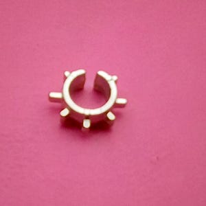 May include: A gold-coloured ear cuff with a sunburst design, featuring a circular shape with multiple outward-pointing spikes. The ear cuff is set against a solid pink background.