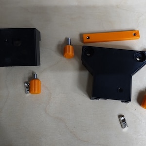 May include: A set of black plastic parts for a 3D printer, including a base, a bracket, and a small box. The set also includes four orange knobs and two silver brackets.