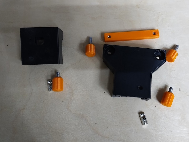 May include: A set of black plastic parts for a 3D printer, including a base, a bracket, and a small box. The set also includes four orange knobs and two silver brackets.