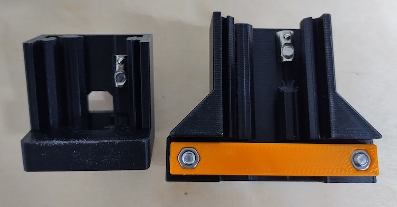 May include: Two black 3D printed parts with a small orange piece in between. The orange piece has two silver screws on either side.