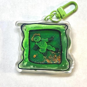 Könnte beinhalten: Ein grüner Acryl-Schlüsselanhänger mit einer Cartoonfigur mit Hut und Handschuhen, eingebettet in einen grünen Rahmen. Der Hintergrund hat Glitzerakzente. Ein grüner Verschluss ist oben befestigt.
