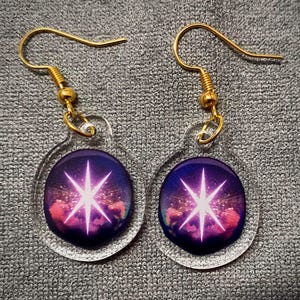 May include: A pair of clear acrylic earrings with gold-colored hooks. Each earring features a circular design with a purple and pink nebula background and a bright white starburst in the center.